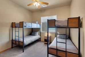 A room with two bunk beds, a ceiling fan, a window with blinds, and a bedside table with a lamp.