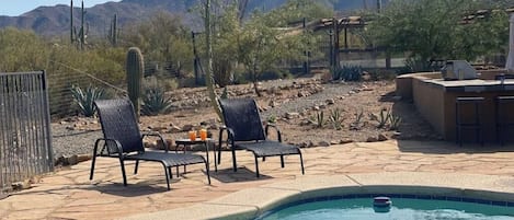 Zona de piscina con sillas y una parrilla, rodeada de vegetación desértica y montañas.