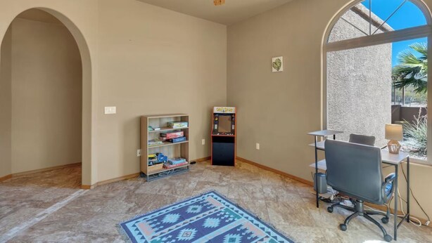Quarto com ventilador de teto, estante, máquina de jogos antiga, escrivaninha com cadeira e uma grande janela com vista para palmeiras.