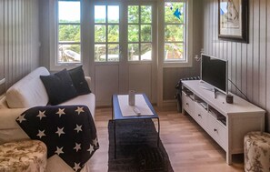Ein gemütliches Wohnzimmer mit einer Couch, einem Couchtisch, einem Fernseher und Blick auf Grün hinter den Fenstern.