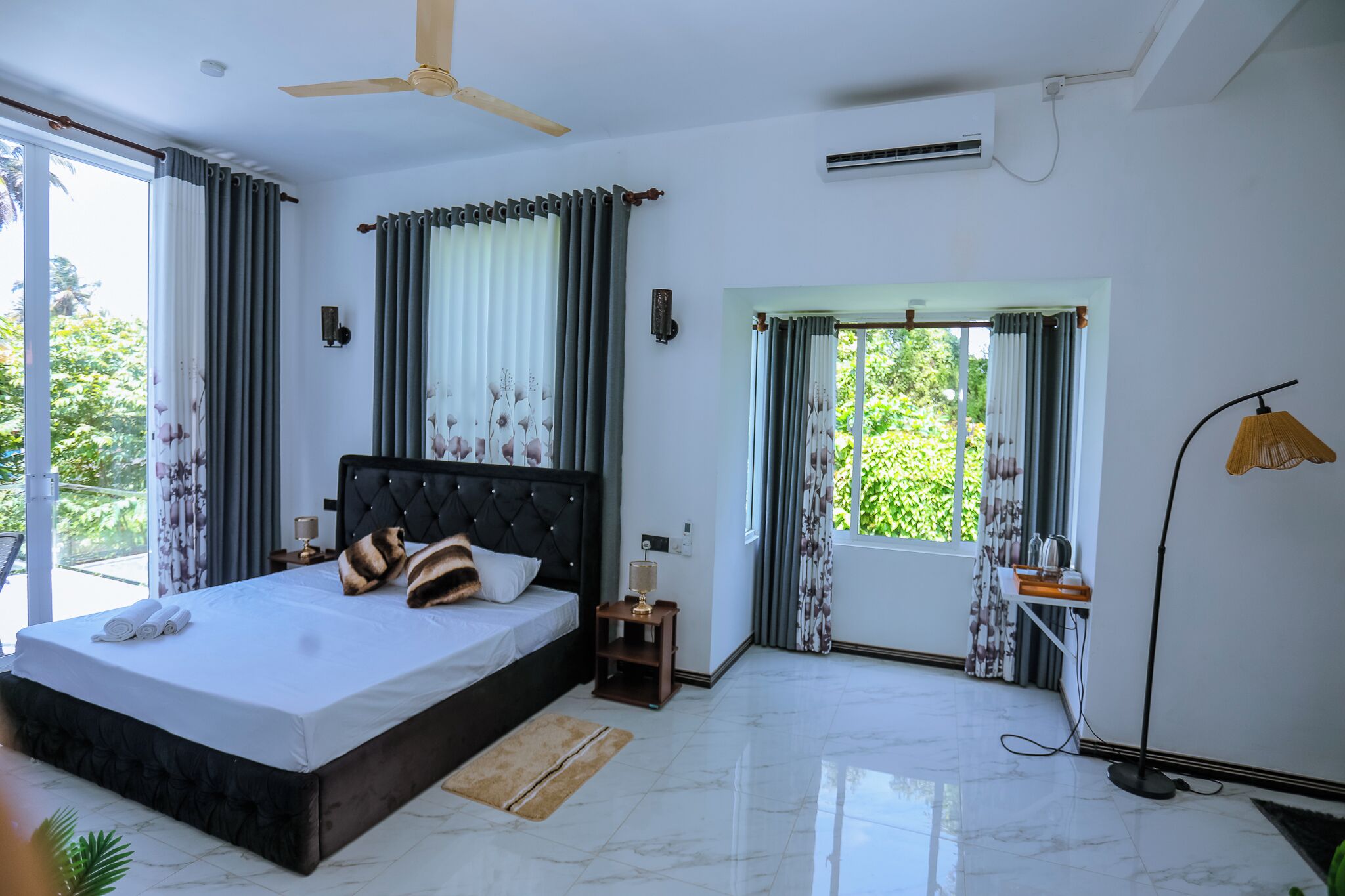 A modern bedroom with a large bed, a ceiling fan, an air conditioning unit, and a view of greenery outside.
