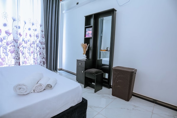 A hotel room with a bed, a desk, a mirror, a trash bin, and a window with curtains.