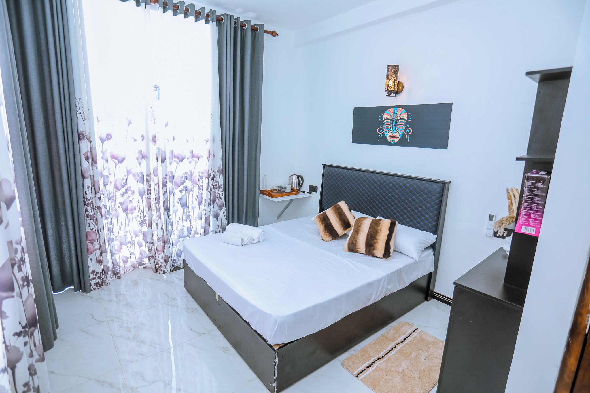 Een hotelkamer met een bed, nachtkastje, wanddecoratie en een raam met gordijnen.
