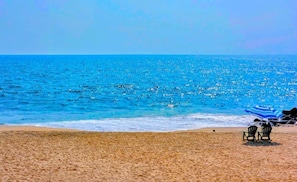 A beach with clear blue water, a sandy shore, and two chairs under a striped umbrella.