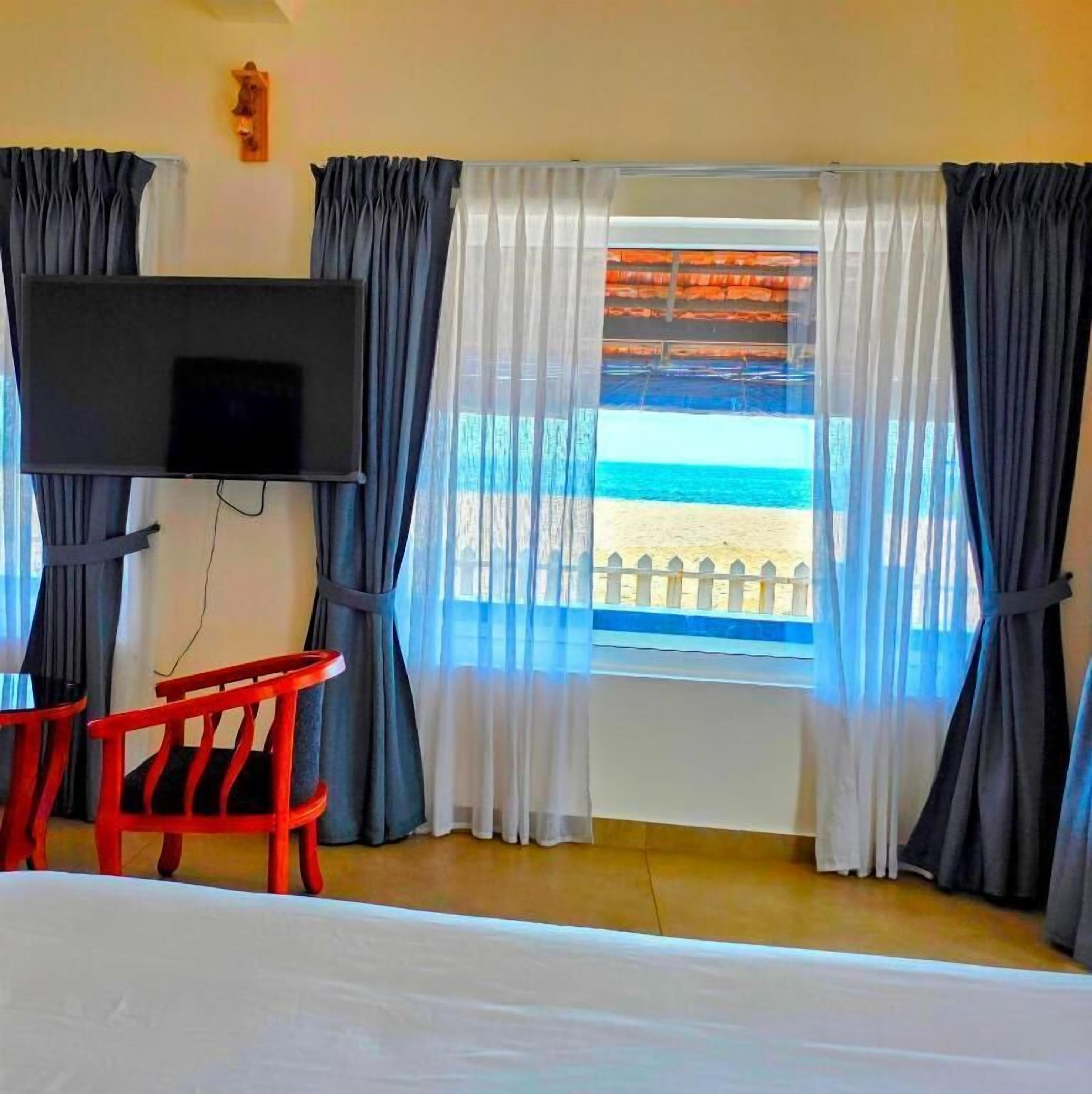 Una habitación con una cama, un televisor montado en la pared, una ventana con vista a la playa y una silla roja.