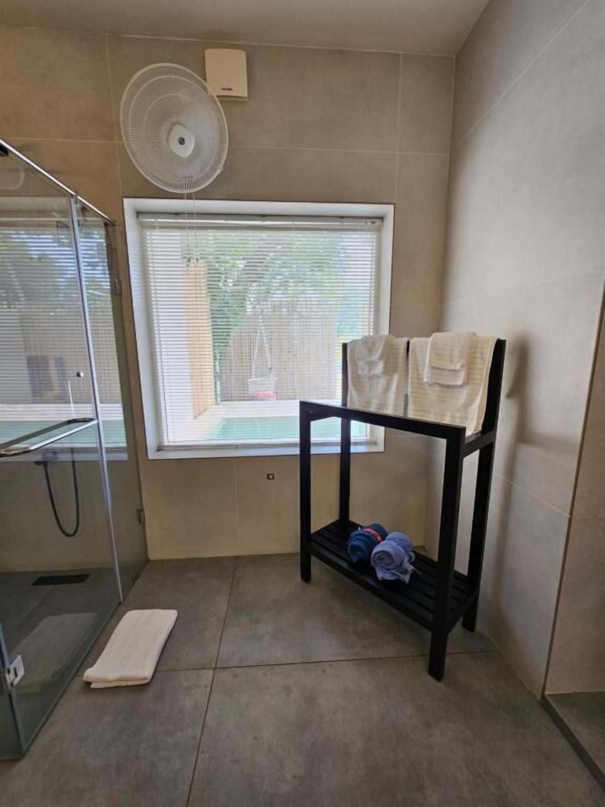 Un baño con cabina de ducha de vidrio, un perchero con toallas, un ventilador y una ventana con persianas.