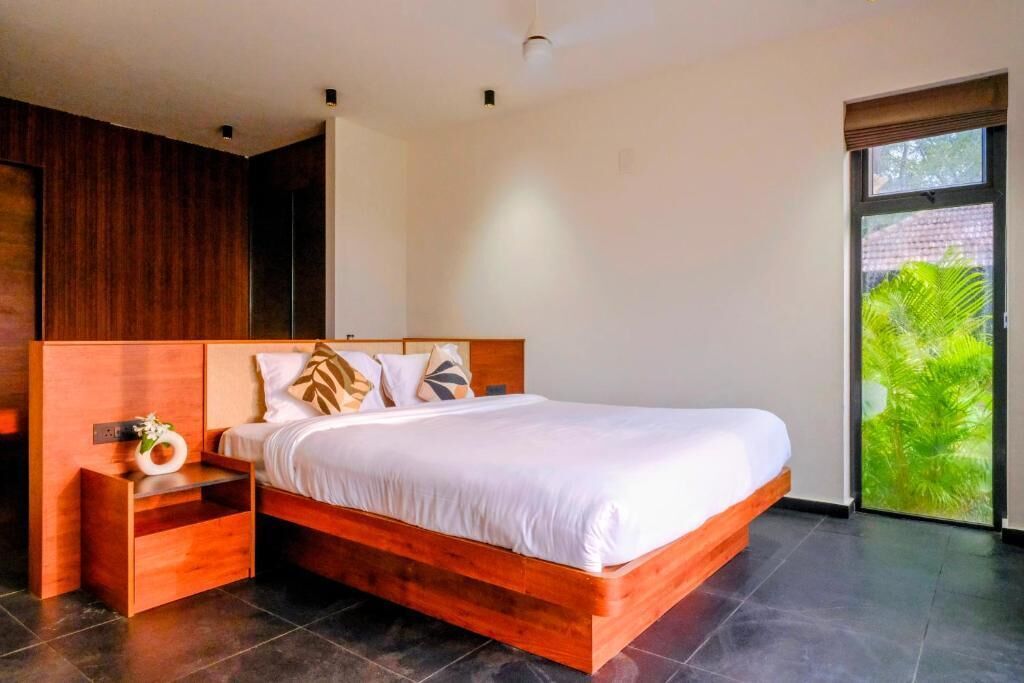 A modern bedroom with a wooden bed frame, a nightstand, and a window with a view of greenery.