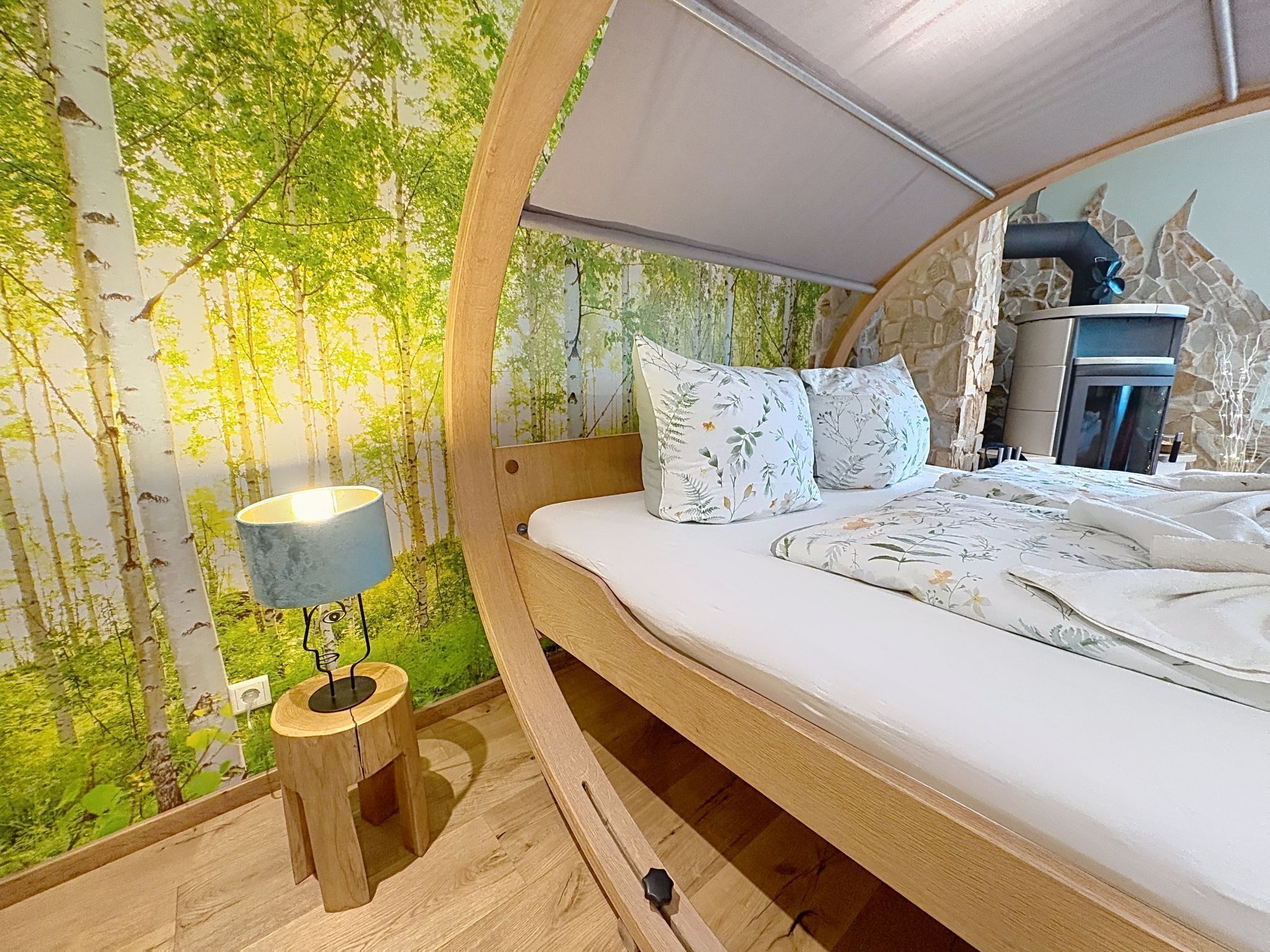 Een gebogen bed met hoofdeinde, een nachtkastje met lamp, en een wand met een bosdecoratie.