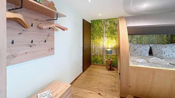 Ein Schlafzimmer mit einem hölzernen Bett, einem an der Wand befestigten Holzregal, einem kleinen Tisch mit einer Lampe und einer Tür zu einem anderen Raum.