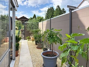 Ein schmaler Pfad führt zu einem Gebäude, im Hintergrund ist eine Holzhütte zu sehen. Überall stehen Kübelpflanzen und der Boden ist mit Kies bedeckt.