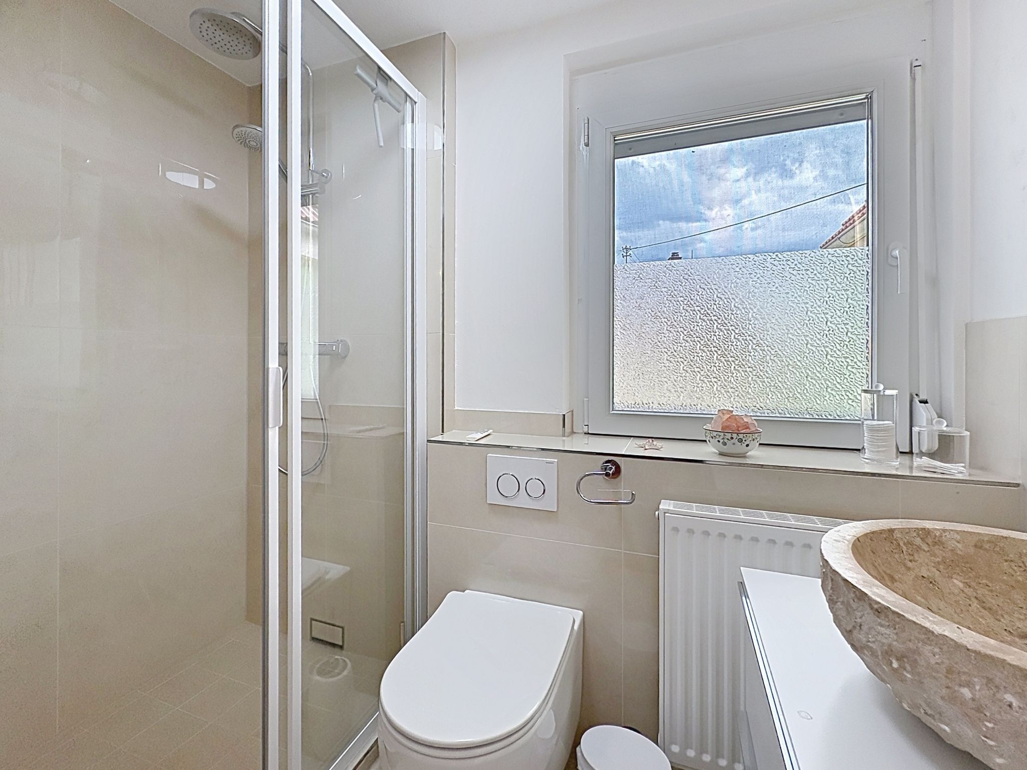 Un baño moderno con cabina de ducha de vidrio, inodoro, lavamanos de piedra y ventana con vistas al exterior.
