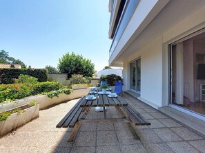 Una terraza en la azotea con una mesa de madera preparada para comer, rodeada de plantas y un edificio con grandes ventanales.