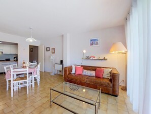 A living room with a glass coffee table, a brown sofa with cushions, a dining area with a white table and chairs, a white fan, and a lamp. - La Frégate by Interhome (Vaux Sur Mer)