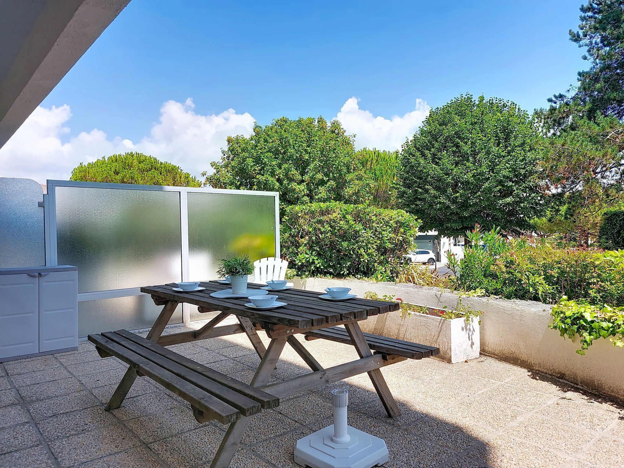 Une terrasse extérieure avec une table et des bancs en bois, entourée de verdure et sous un ciel dégagé.