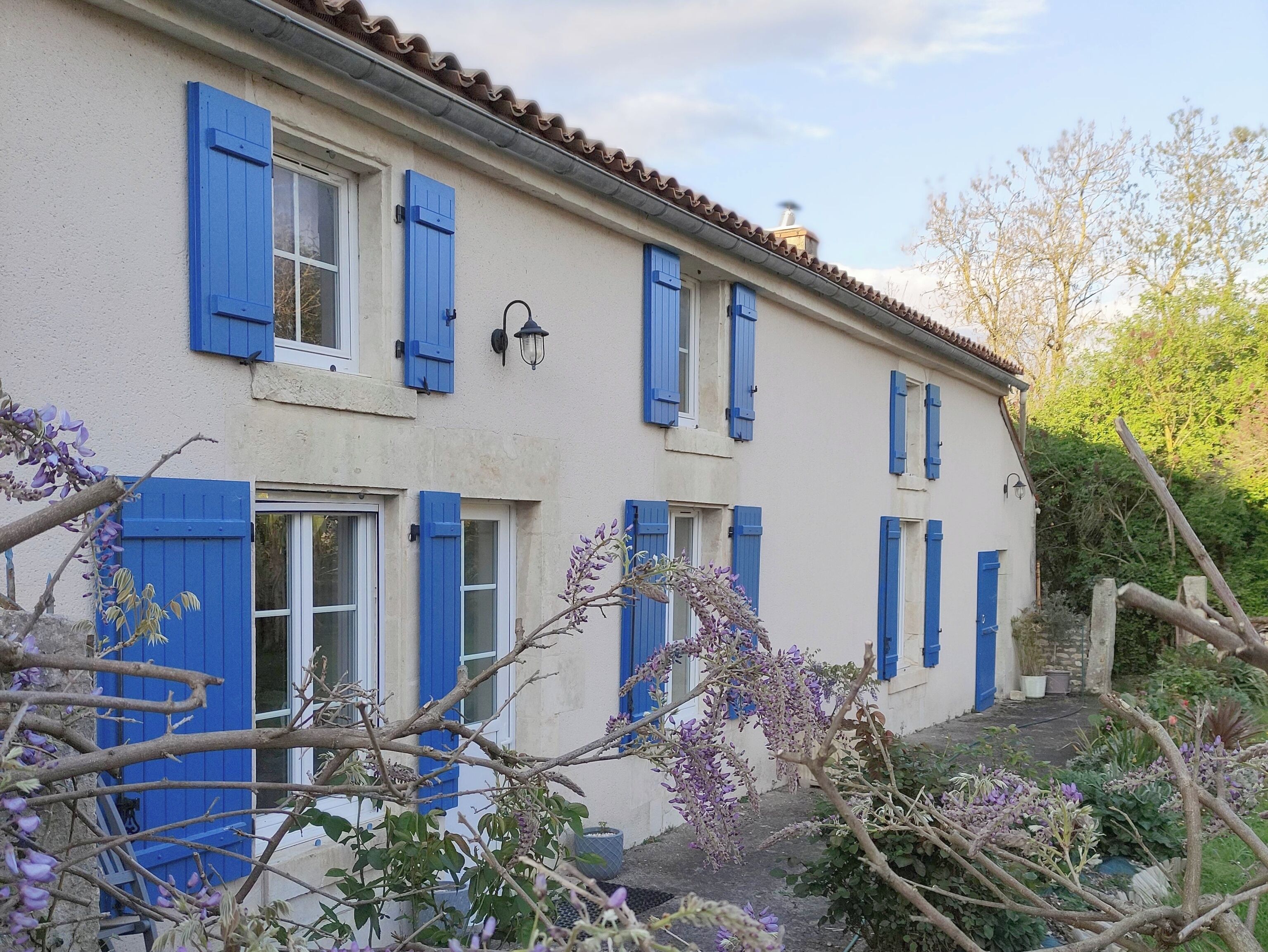 A two-story building with blue shutters, a tiled roof, and a garden with purple flowers.