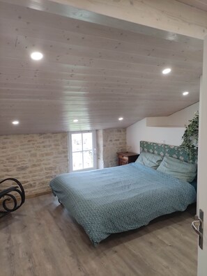 A bedroom with a sloped ceiling, a bed with patterned bedding, a nightstand, and a window. - Les Glycines - A Large, Charming Vacation Rental in the Heart of the Marais Poitevin (Saint-Sigismond)