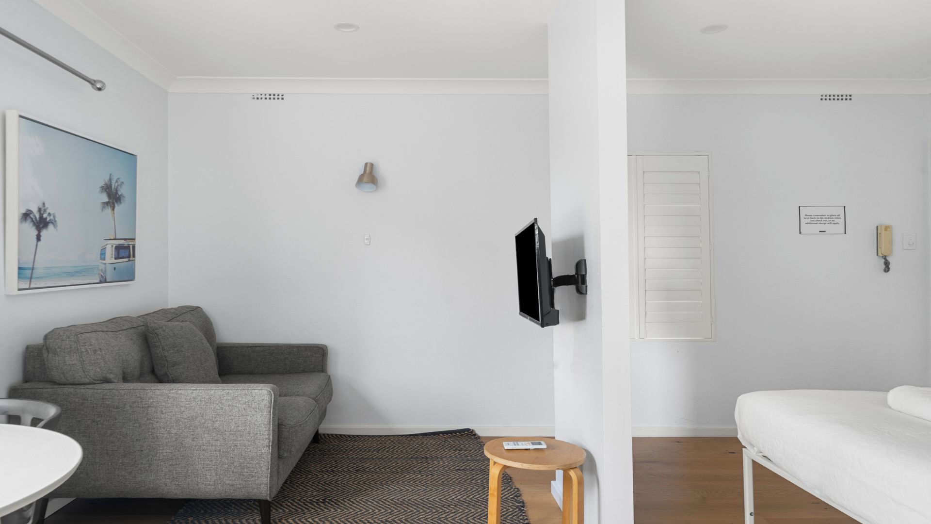 Ein modernes, minimalistisches Zimmer mit grauem Sofa, einem kleinen runden Tisch, einem an der Wand montierten Fernseher und einem weißen Bett.