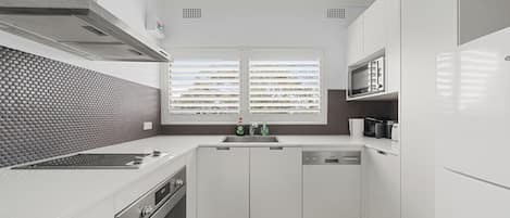 A modern kitchen with white cabinetry, a stainless steel range hood, a built-in microwave, and a window with blinds.