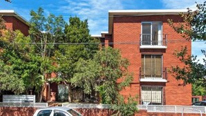 Un edificio de ladrillo de dos pisos con un balcón en el segundo nivel, rodeado de árboles y una cerca blanca.