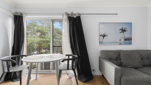 Una habitación con un sofá gris, una mesa redonda con sillas, un ventanal con cortinas y un cuadro de una playa.