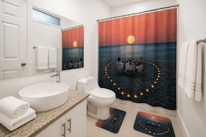Ein Badezimmer mit einem großen Waschbecken, Toilette und einer Duschkabine, deren Vorhang einen Sonnenuntergang über Wasser zeigt.