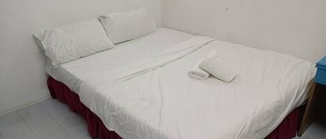 A single bed with a white mattress and red skirted base, a white pillow, and a folded white towel.