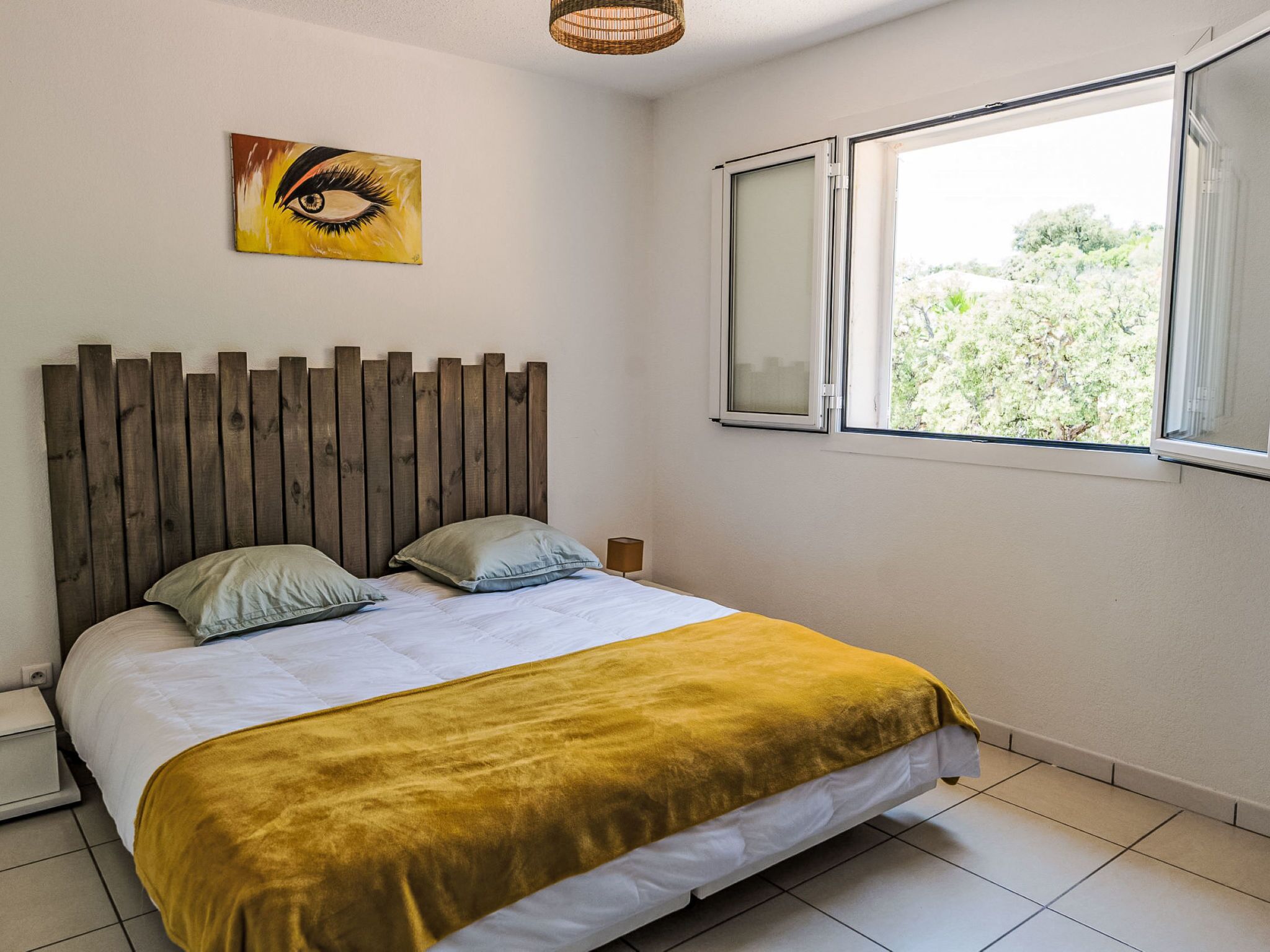 A bedroom with a wooden headboard, a bed with white and yellow bedding, a nightstand, a lamp, and a window with a view of greenery.