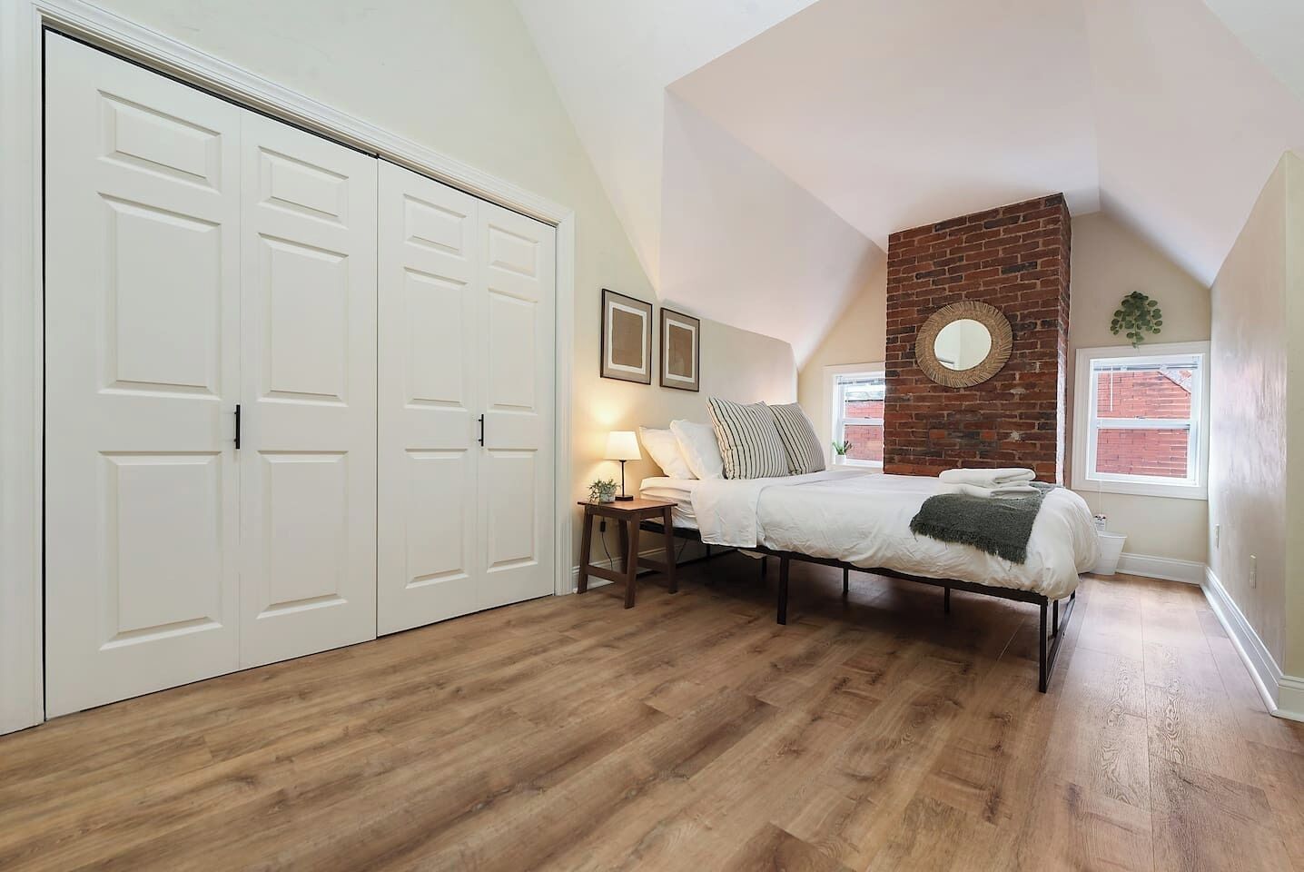 A bedroom with a bed, a nightstand, a lamp, a brick accent wall, and a large closet.