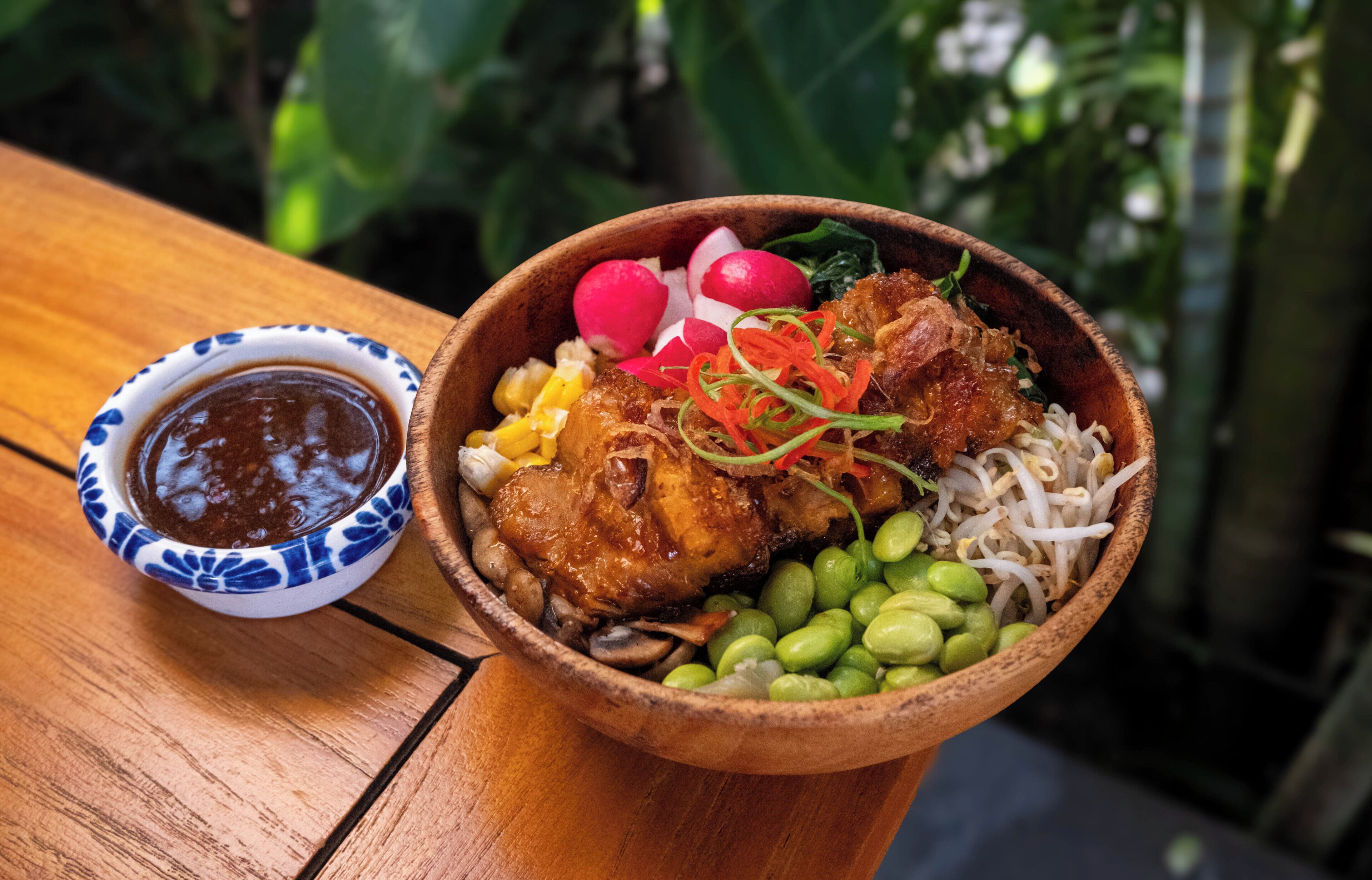 A wooden table with a bowl of food containing meat, vegetables, and a small bowl of sauce.