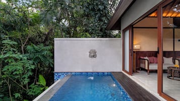 A swimming pool with a waterfall feature, surrounded by lush greenery and a modern building with a glass door leading to a seating area.