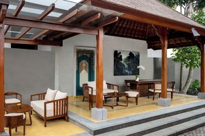 Uma área de estar coberta ao ar livre, com móveis de madeira, uma porta com design elaborado e uma pintura de uma cascata.