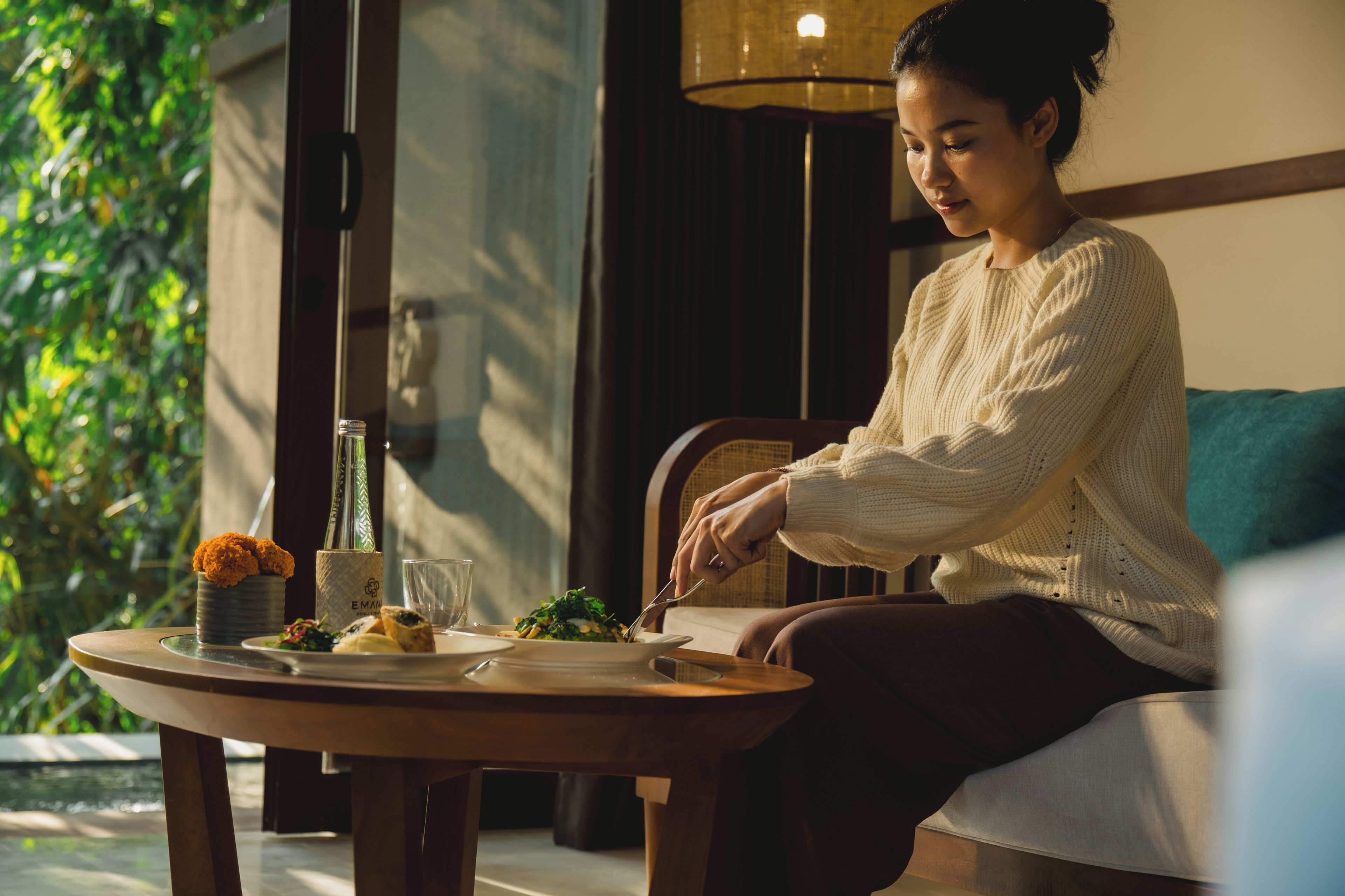 A person seated at a table with a meal, a lamp, and a view of greenery outside.