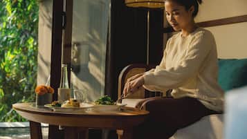 A person seated at a table with a meal, a lamp, and a view of greenery outside.