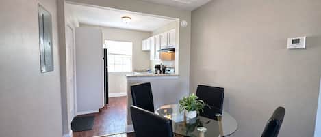 A dining area with a glass table and black chairs, a kitchen in the background, and a thermostat on the wall.