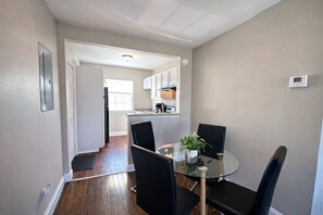 A dining area with a glass table and black chairs, a kitchen in the background, and a thermostat on the wall.