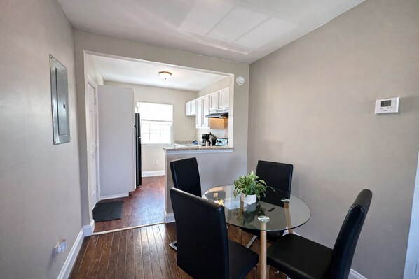 A dining area with a glass table and black chairs, a kitchen in the background, and a thermostat on the wall.