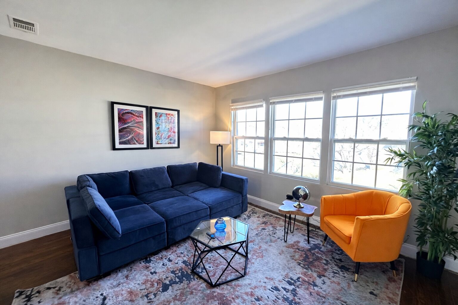 Un salon avec un canapé bleu, un fauteuil orange, une table basse et une grande fenêtre avec des stores.