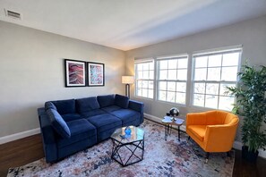 A living room with a blue sofa, an orange armchair, a coffee table, and a large window with blinds.