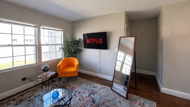Ein Zimmer mit großem Fenster, einer Pflanze, einem an der Wand befestigten Fernseher, einem Spiegel und einem kleinen Tisch mit Lampe.