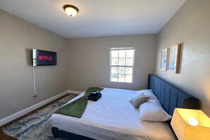 Ein Schlafzimmer mit einem Bett, einem an der Wand montierten Fernseher, einem Fenster und einer Nachttischlampe.
