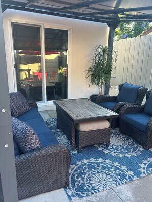 Ein Terrassenbereich im Freien mit Korbmöbeln, einem gemusterten Teppich und einer Glasschiebetür, die auf eine Terrasse mit Tisch und Stühlen führt.