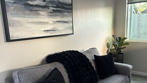 A grey sofa with black cushions, a large abstract painting, an air conditioning unit, a window with a view, and a potted plant. - Cozy Casita in the Valley (Lemon Grove)