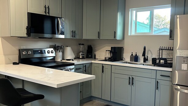 A modern kitchen with grey cabinets, a central island with bar stools, a stainless steel refrigerator, and a window providing natural light. - Cozy Casita in the Valley (Lemon Grove)