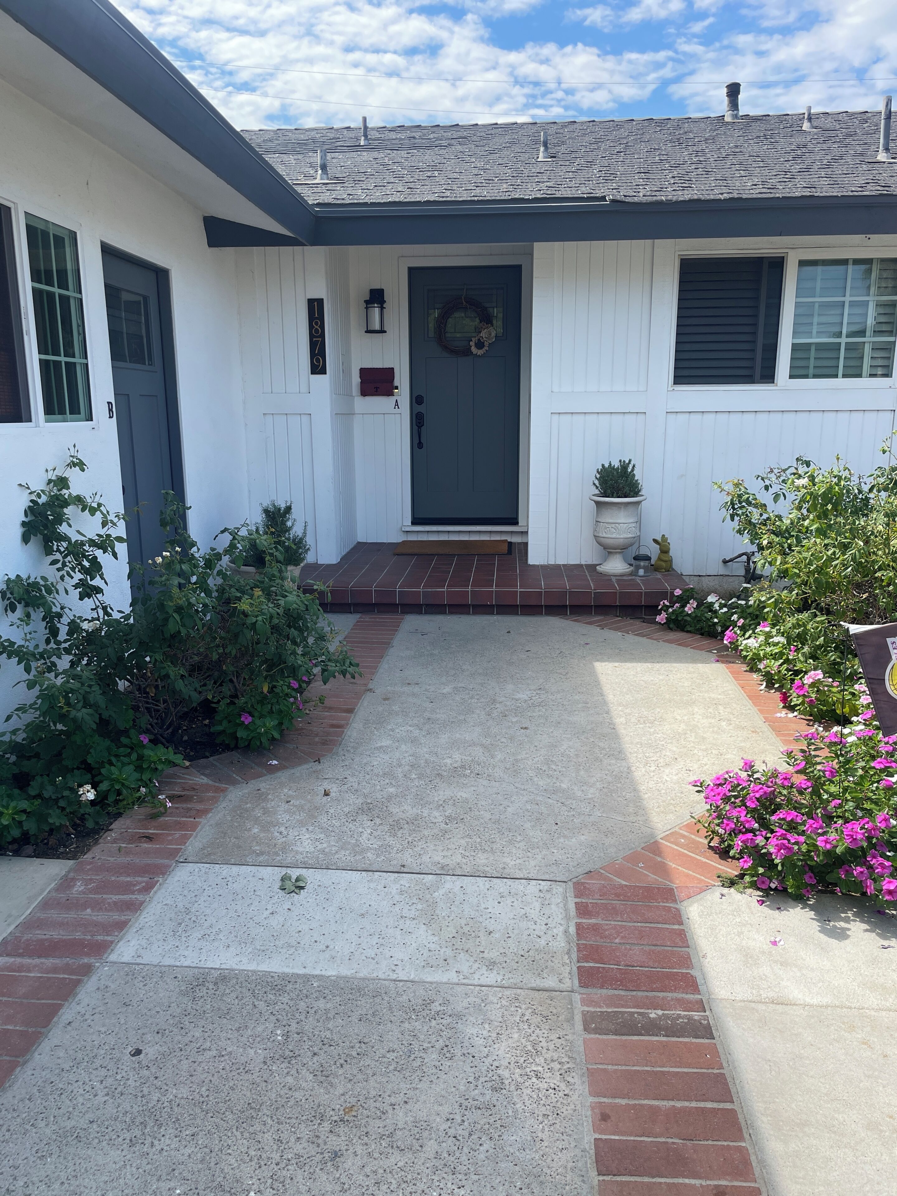 A white house with a dark front door, a brick pathway leading to the entrance, and a small garden with flowers.