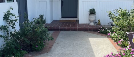 A white house with a dark front door, a brick pathway leading to the entrance, and a small garden with flowers.