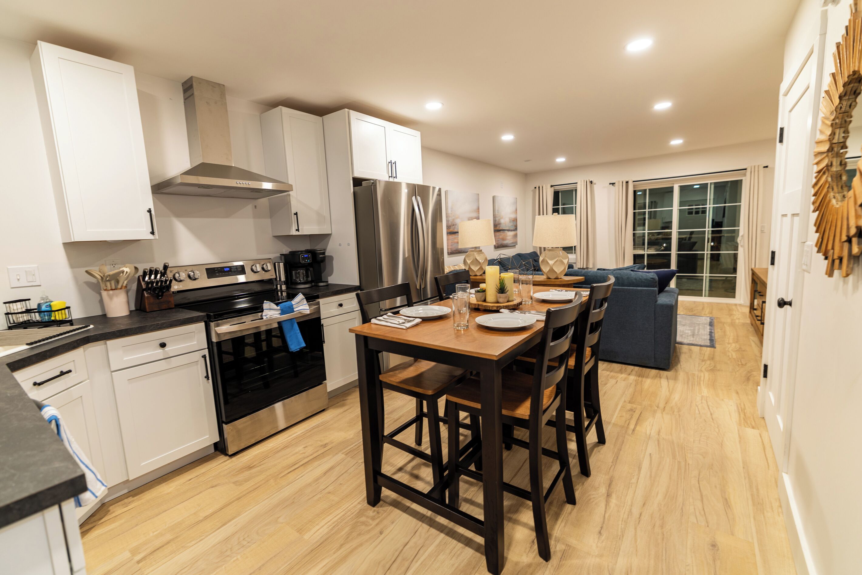 A modern kitchen with a dining area, wooden flooring, and a view of the outdoors through a sliding glass door.