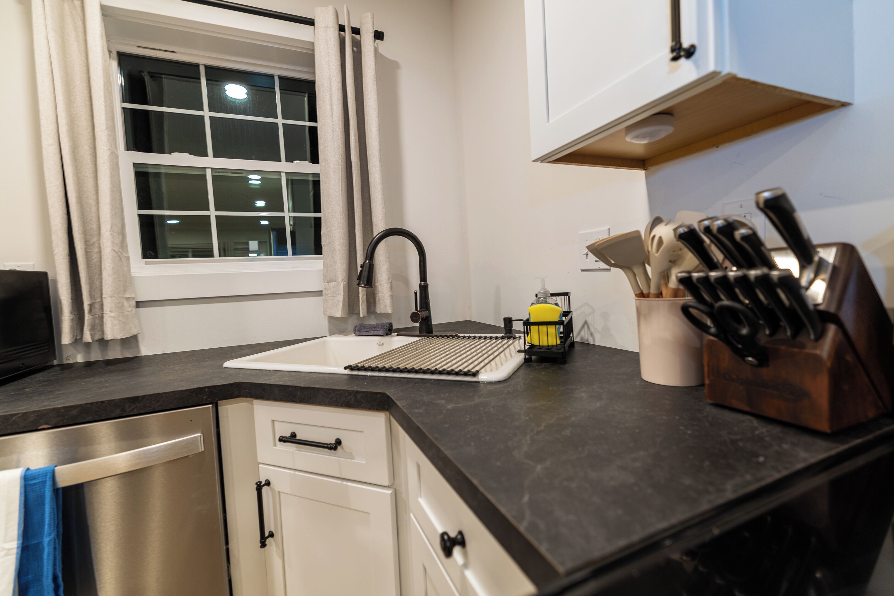 Una cocina con un lavamanos de acero inoxidable, un bloque de cuchillos y una ventana con cortinas.