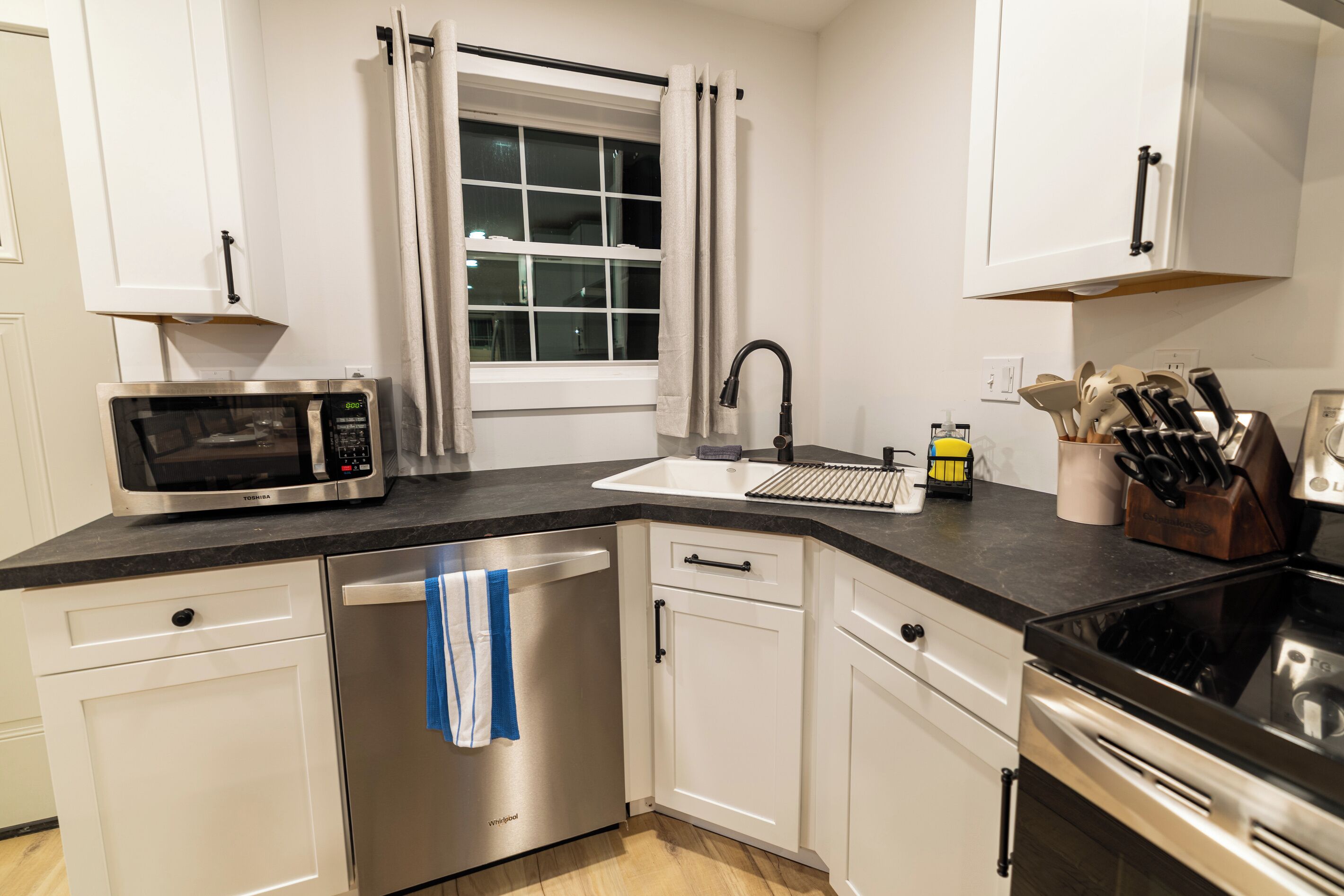 A modern kitchen with white cabinetry, a stainless steel dishwasher, a microwave, a sink with a faucet, and a stove.