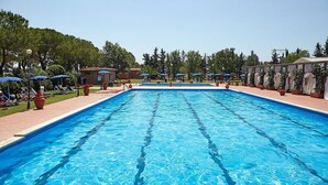 Piscina rectangular con agua azul cristalina, rodeada de sillas de descanso y sombrillas, con árboles y un edificio al fondo.