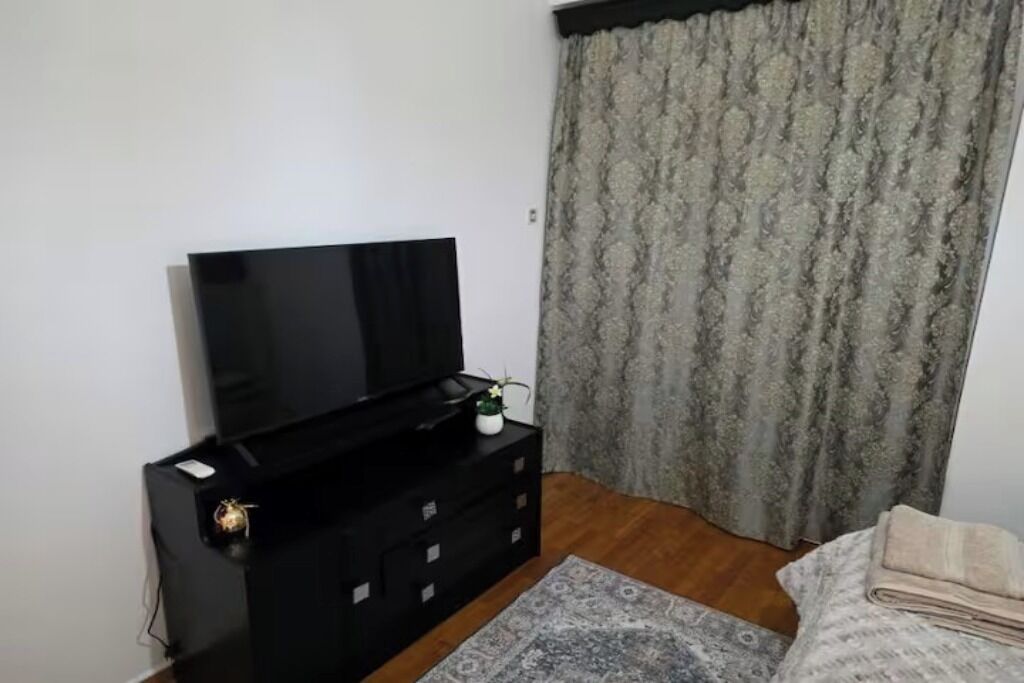 A room with a television on a black stand, a patterned rug, a bed with a folded blanket, and a curtain with a floral pattern.